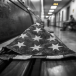 A folded American flag resting on a wooden VA hospital waiting-room bench, black and white with a gold accent.