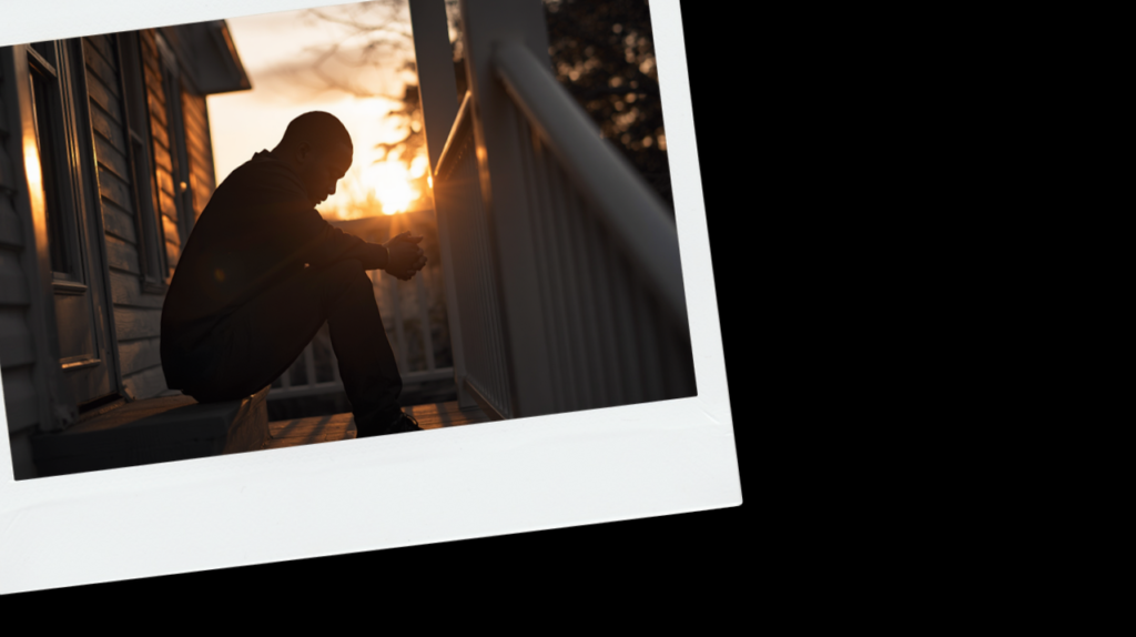 A Black man sitting alone on porch steps at golden hour, black and white with a warm gold rim light.