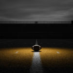 An empty college football stadium at dusk with a single spotlight on a football at midfield, black and white with gold light.