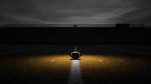 An empty college football stadium at dusk with a single spotlight on a football at midfield, black and white with gold light.